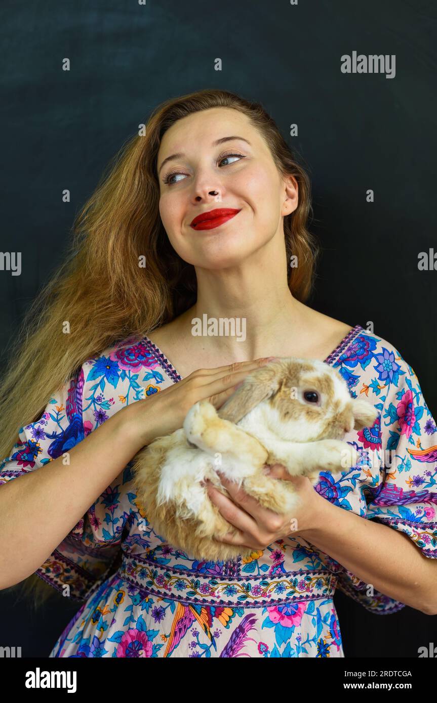 Eine wunderschöne russin mit langen blonden Haaren, die ein Kaninchentier in der Hand an die schwarze Wand hält Stockfoto