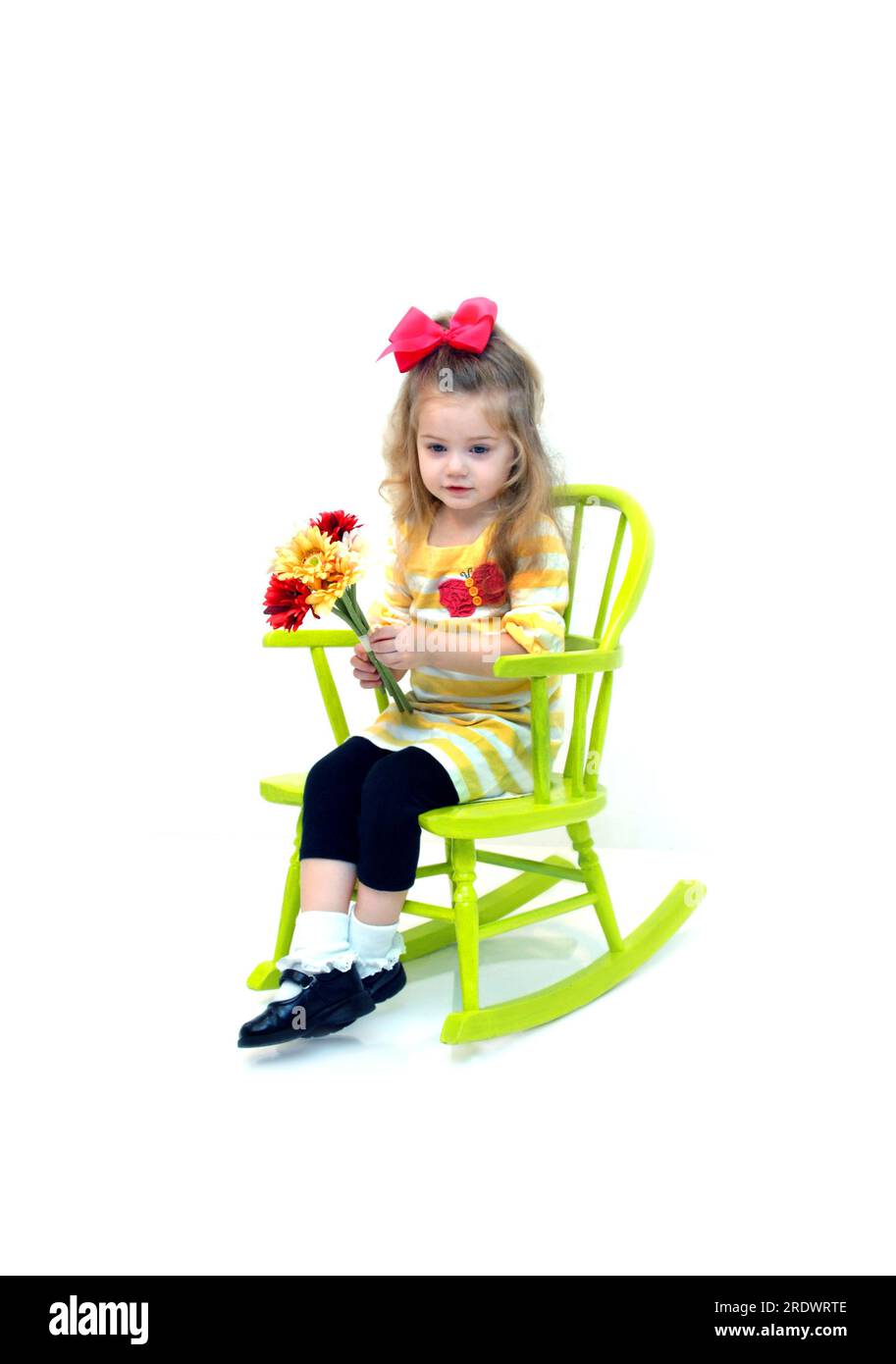 Das kleine Mädchen sitzt in einem hellgrünen, bemalten Schaukelstuhl aus Holz. Sie hält einen Blumenstrauß in der Hand. Stockfoto