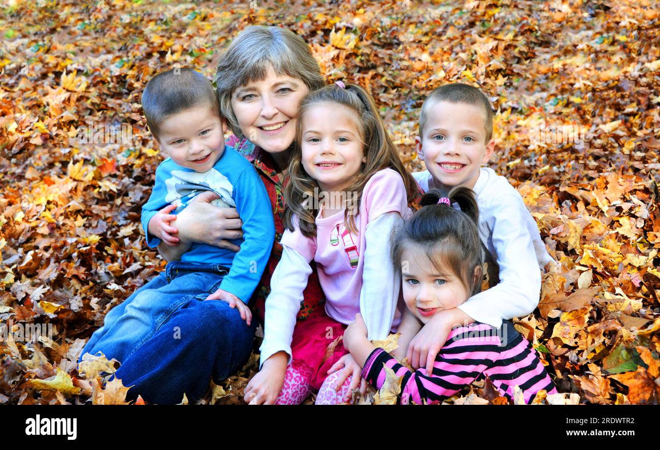 Als sie Herbstblätter harkte, hielt diese Großmutter an, um all ihre Enkel zu umarmen. Sie sitzen auf einem Laubhaufen und lächeln glücklich. Die Kinder einschl Stockfoto
