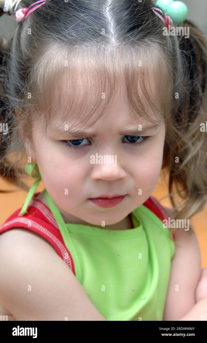 Das kleine Mädchen lässt ihre Gefühle mit seiner zerklüfteten Stirn und den gekreuzten Armen zeigen. Sie hat Zöpfe und trägt eine grüne Schürze und ein rotes Hemd. Stockfoto