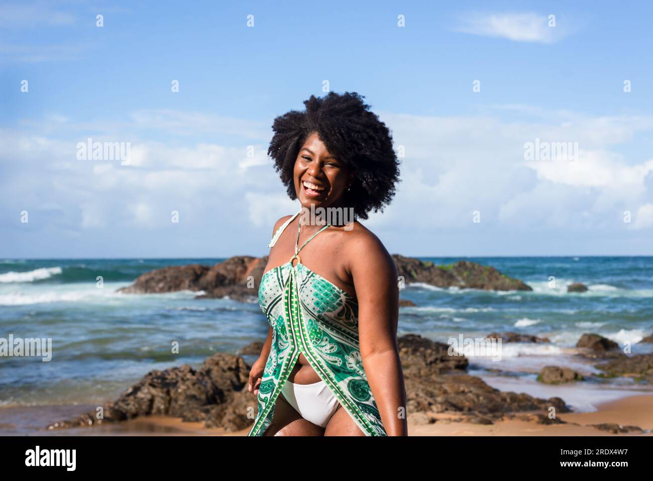 Porträt einer wunderschönen, schwarzhaarigen Frau, die lächelt und glücklich am Strand steht. Im Hintergrund liegen die Wolken auf Felsen und das Meer. Rio Vermelho, Salvad Stockfoto