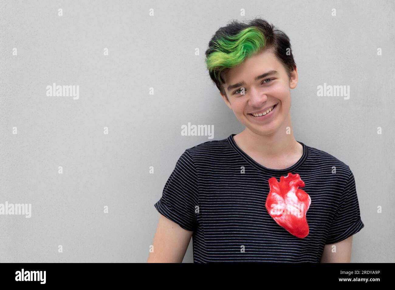 Lächelnder Teenager mit gefärbtem grünen Haar und künstlichem Herz vor grauem Hintergrund Stockfoto