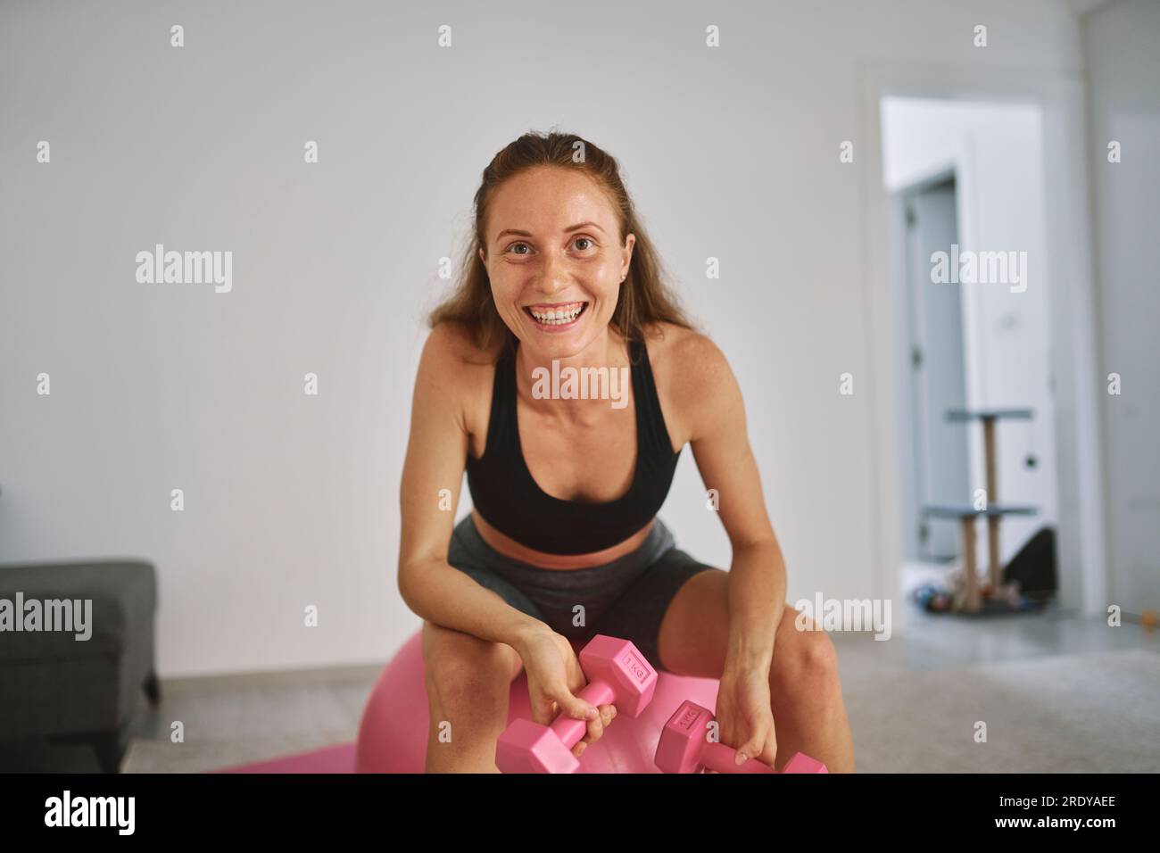 Glückliche junge Frau, die zu Hause mit Kurzhanteln auf einem Fitnessball sitzt Stockfoto