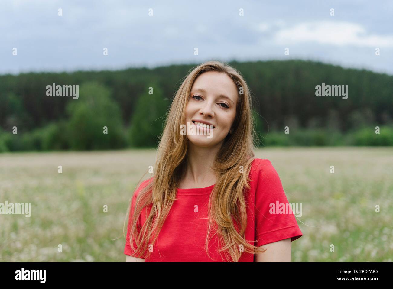 Lächelnde junge Frau, die ein rotes T-Shirt mit blondem Haar auf dem Feld trägt Stockfoto