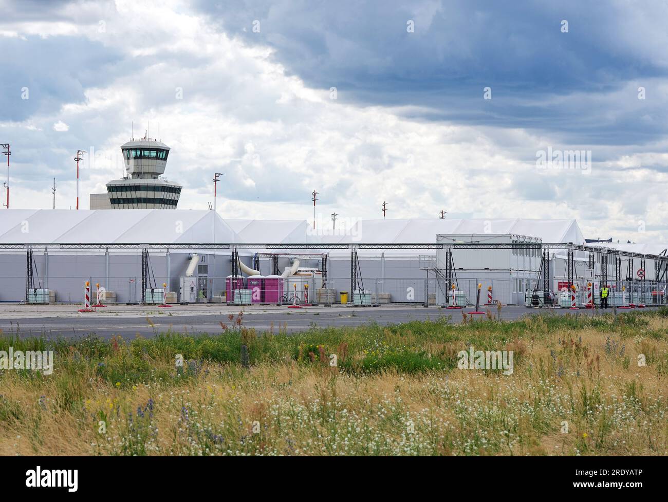 Berlin, Deutschland. 12. Juli 2023. Der Turm des ehemaligen Flughafens Berlin-Tegel hinter den Zelten, in denen derzeit Flüchtlinge untergebracht sind. Auf dem 500 ha großen Gelände befinden sich ein Forschungs- und Industriepark für urbane Technologien, „Berlin TXL - die Urban Tech Republic“, ein Park und das Schumacher-Viertel, ein neues Wohnviertel. Mit der Entwicklung und dem Management wurde das staatliche Unternehmen Tegel Projekt GmbH beauftragt. Kredit: Soeren Stache/dpa/Alamy Live News Stockfoto