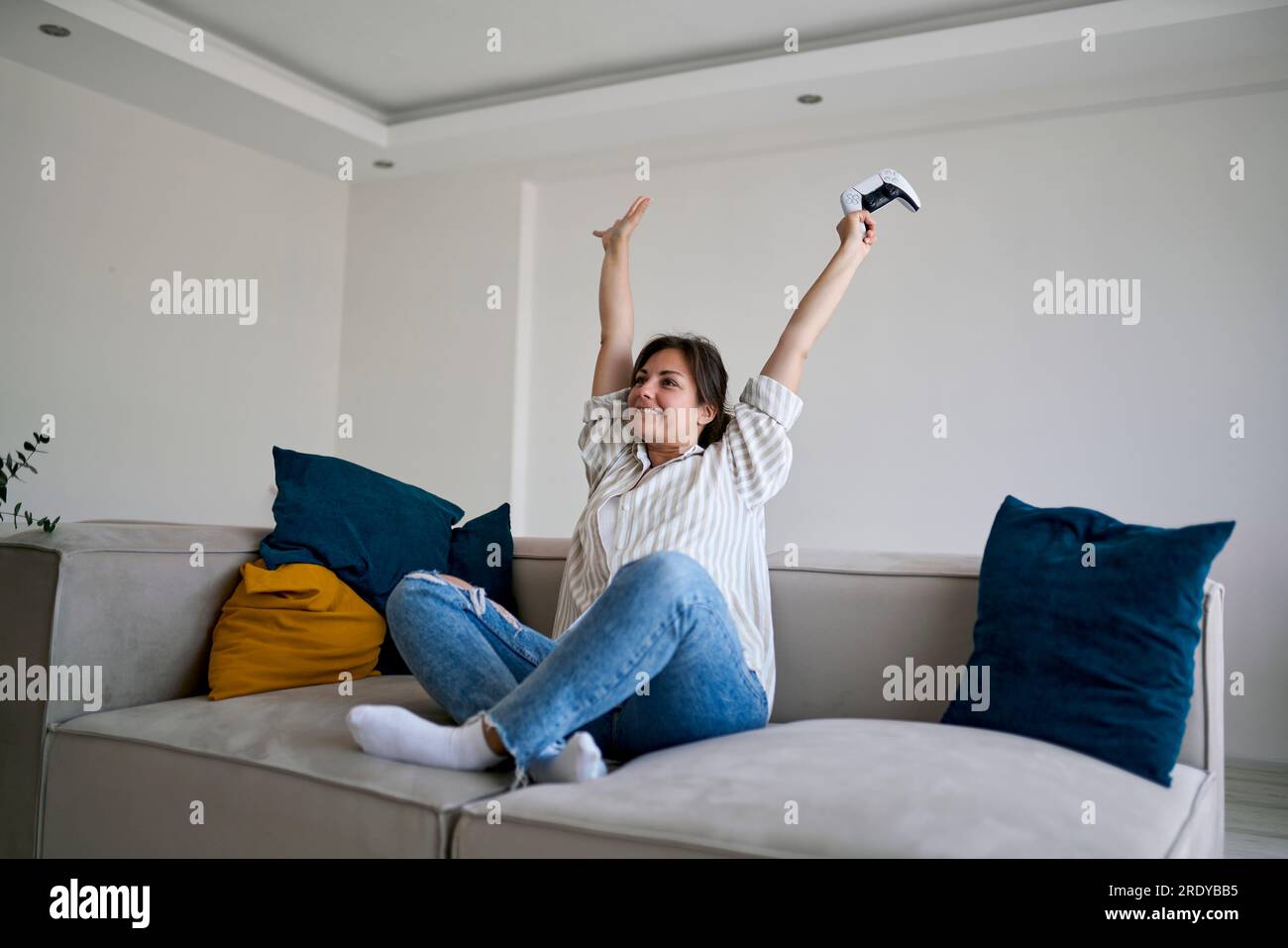 Glückliche Frau mit erhobenen Armen, die zu Hause auf dem Sofa sitzt Stockfoto