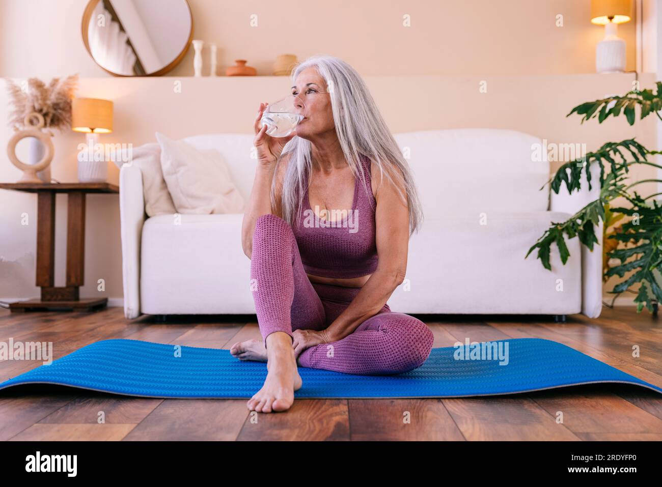 Eine Frau trinkt Wasser und sitzt zu Hause auf einer Yogamatte Stockfoto