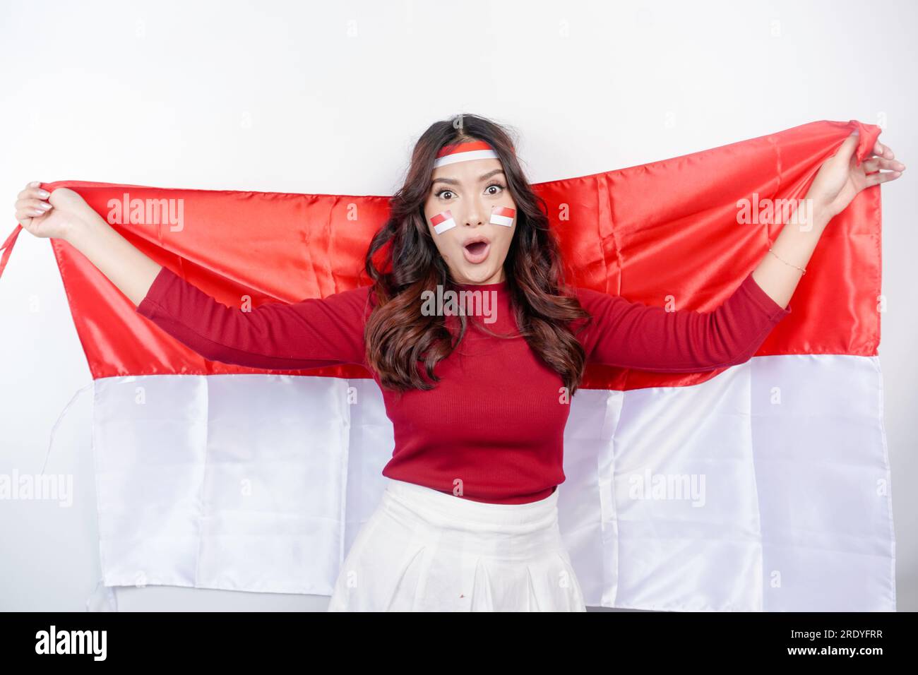 Ein Porträt einer schockierten Asiatin, die ein Kopfband trug, während sie Indonesiens Flagge mit offenem Mund hielt, isoliert von weißem Hintergrund. Indonesien' Stockfoto