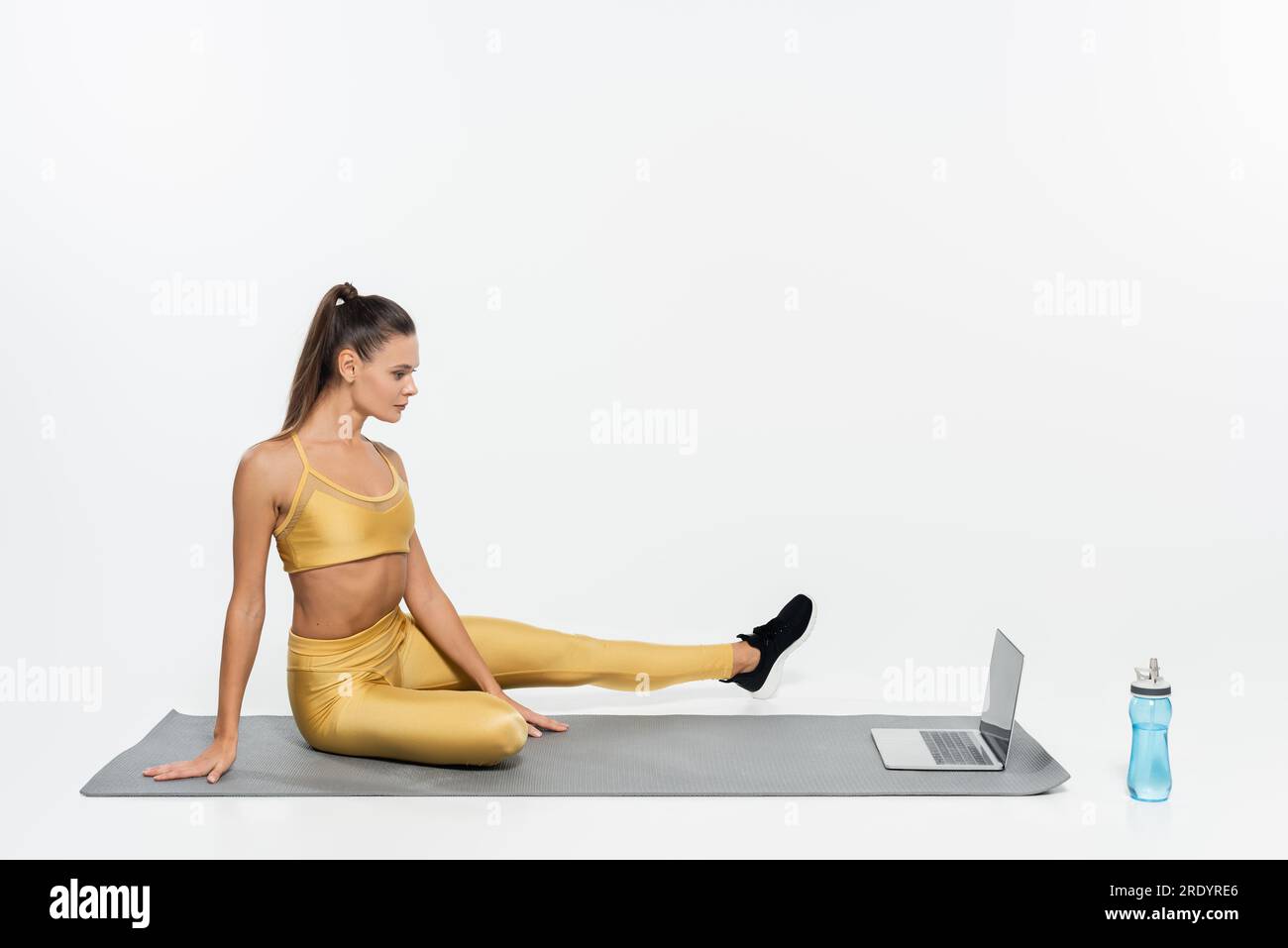 E-Sport, Frau in Sportbekleidung sitzt neben Laptop und Flasche auf Fitnessmatte, weißer Hintergrund Stockfoto