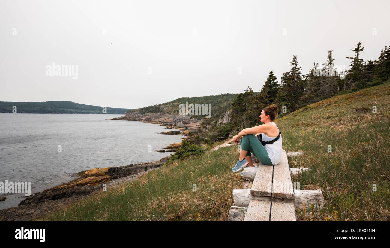 Eine Frau sitzt auf einem hölzernen Pfad und sieht auf einer Wanderung aufs Meer. Stockfoto
