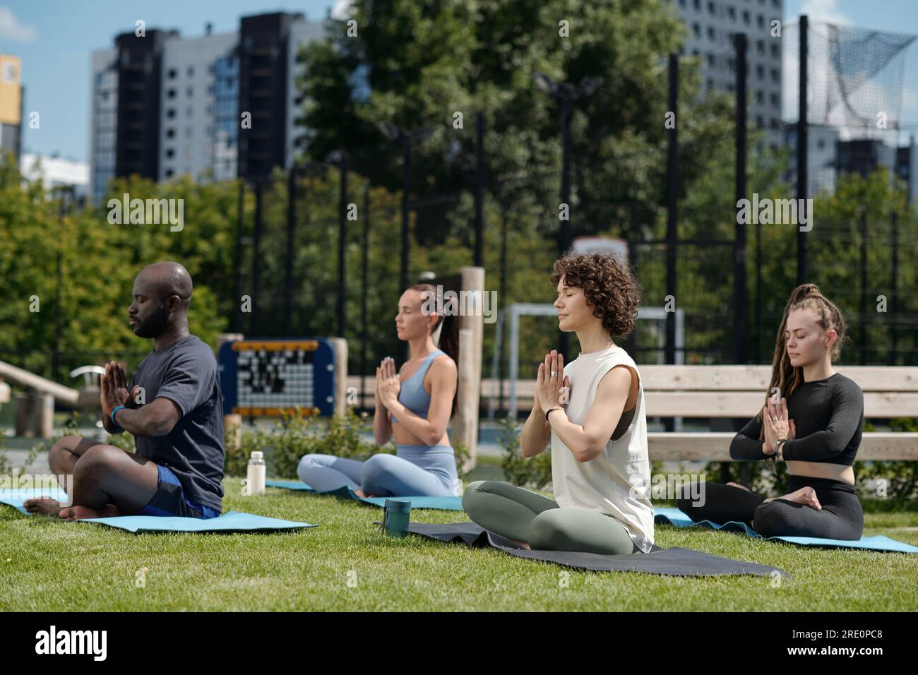 Seitenansicht einer Gruppe interkultureller Athleten, die Yoga im Freien üben, während sie auf Matten sitzen, mit überkreuzten Beinen und Handflächen an der Brust zusammengelegt Stockfoto