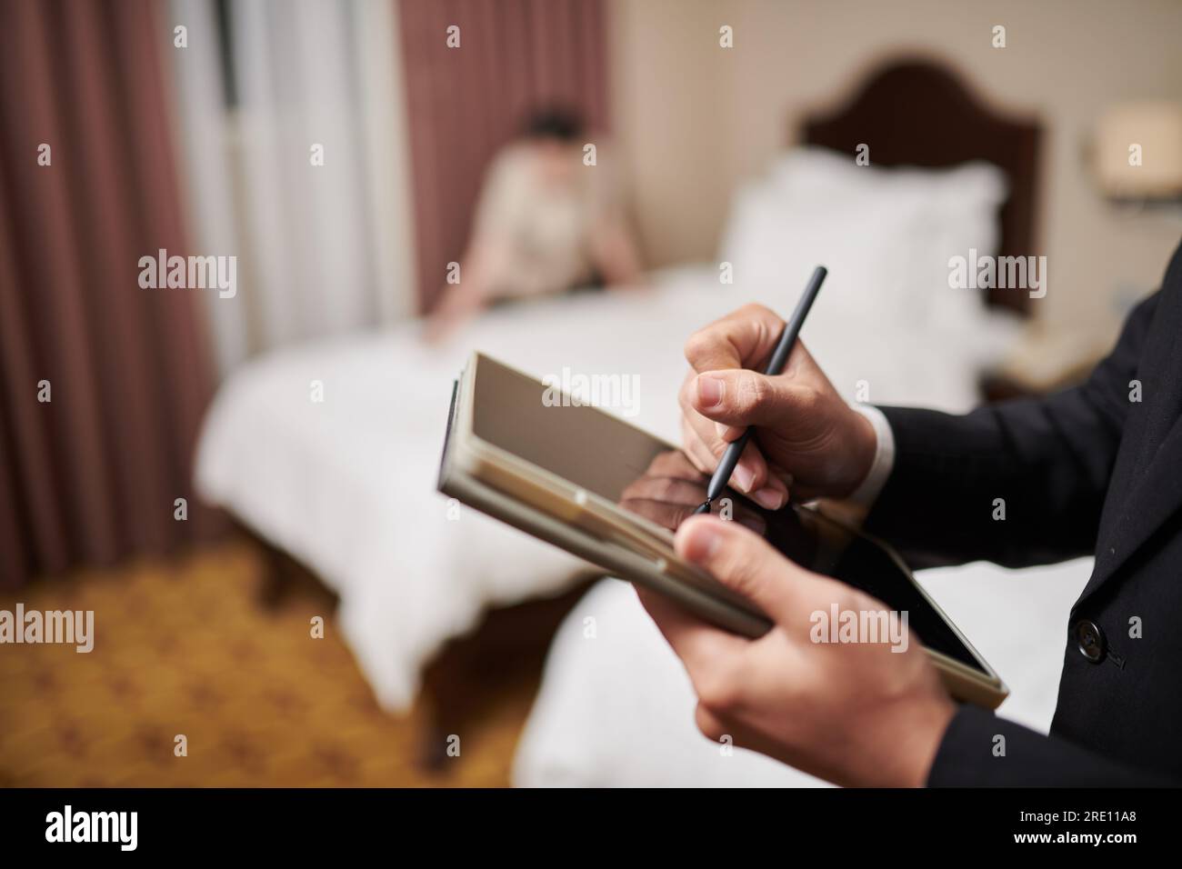 Nahaufnahme des Hotelmanagers, der das Formular auf der Registerkarte ausfüllt, lässt Computer laufen, wenn das Zimmermädchen im Hintergrund das Bett macht Stockfoto