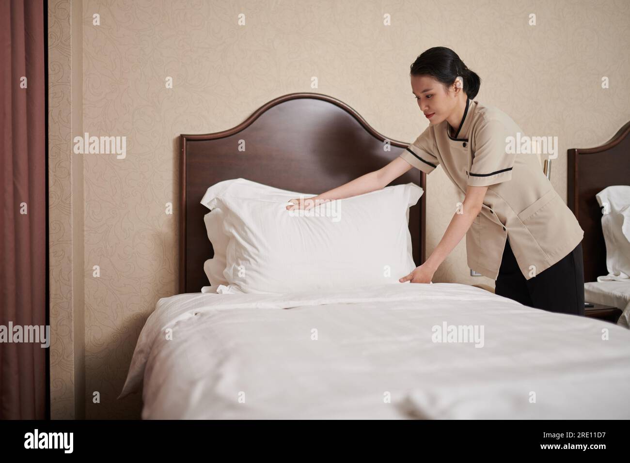 Lächelndes, junges, asiatisches Zimmermädchen, das die Bettwäsche wechselt und das Bett macht Stockfoto