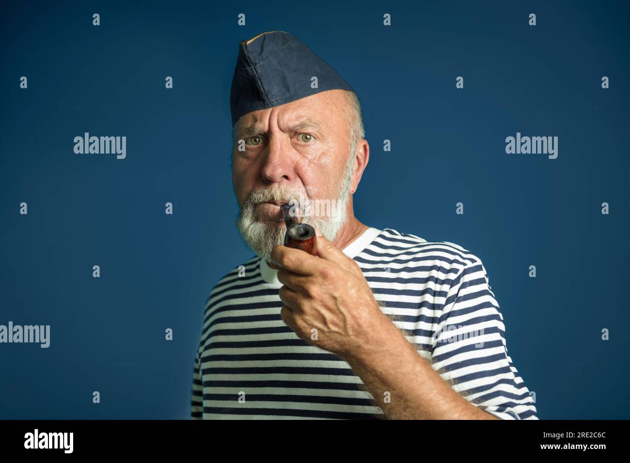 Porträt eines alten Seemanns mit einer Raucherpfeife auf dunkelblauem Hintergrund. In einem Marinehemd mit Streifenmuster und einer blauen Mütze Stockfoto