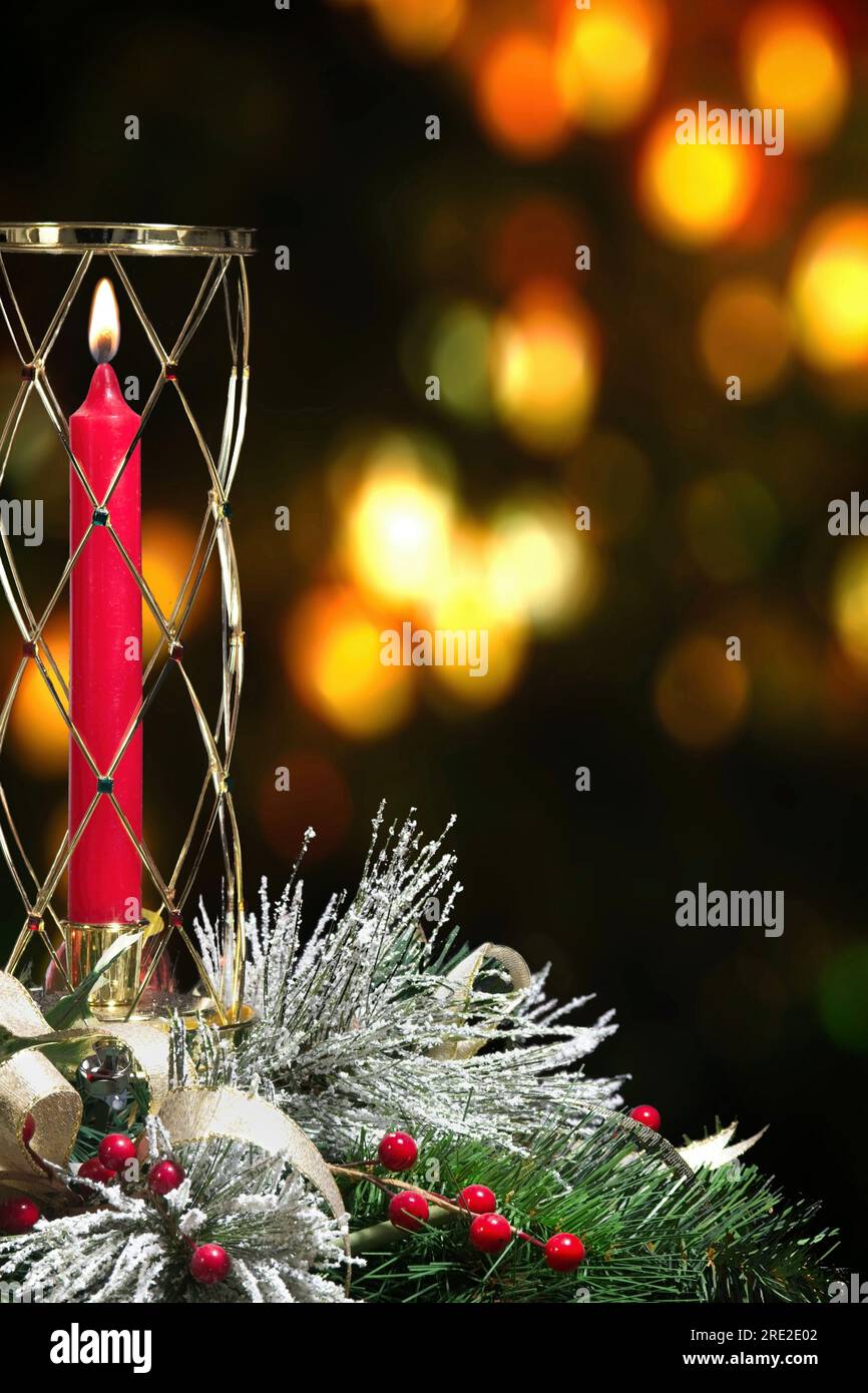 Zur Weihnachtsdekoration gehören Kunstkiefernadeln, Beeren, Schneekornadeln und ein Band, mit einer beleuchteten, langstieligen roten Kerze in einem Glasbehälter. Stockfoto