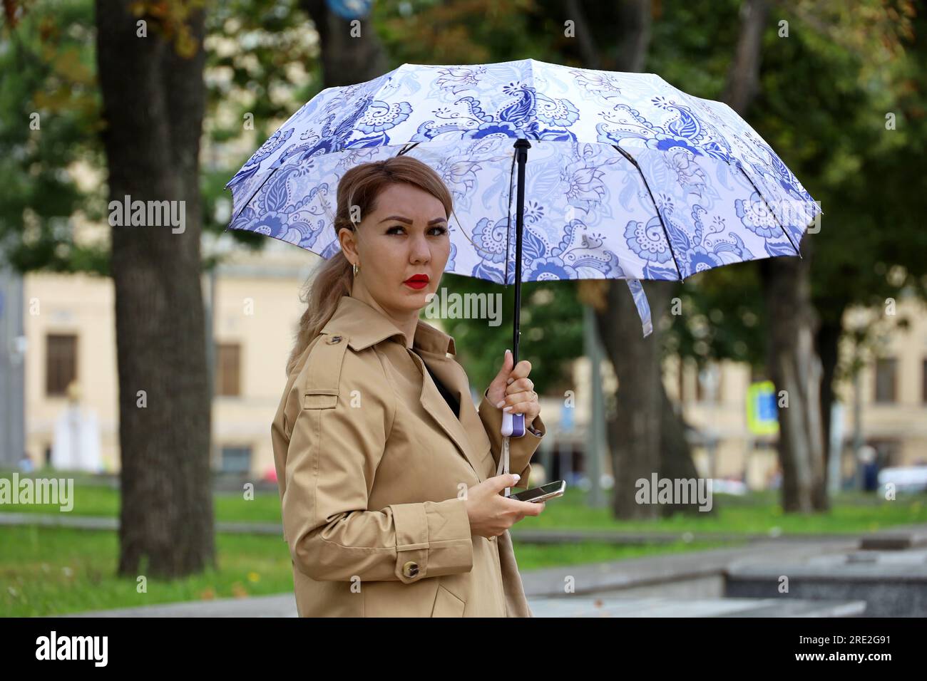 Asiatische Frau mit Schirm und Smartphone, die auf einer Straße steht. Regenwetter in der Stadt, Sommersturm Stockfoto