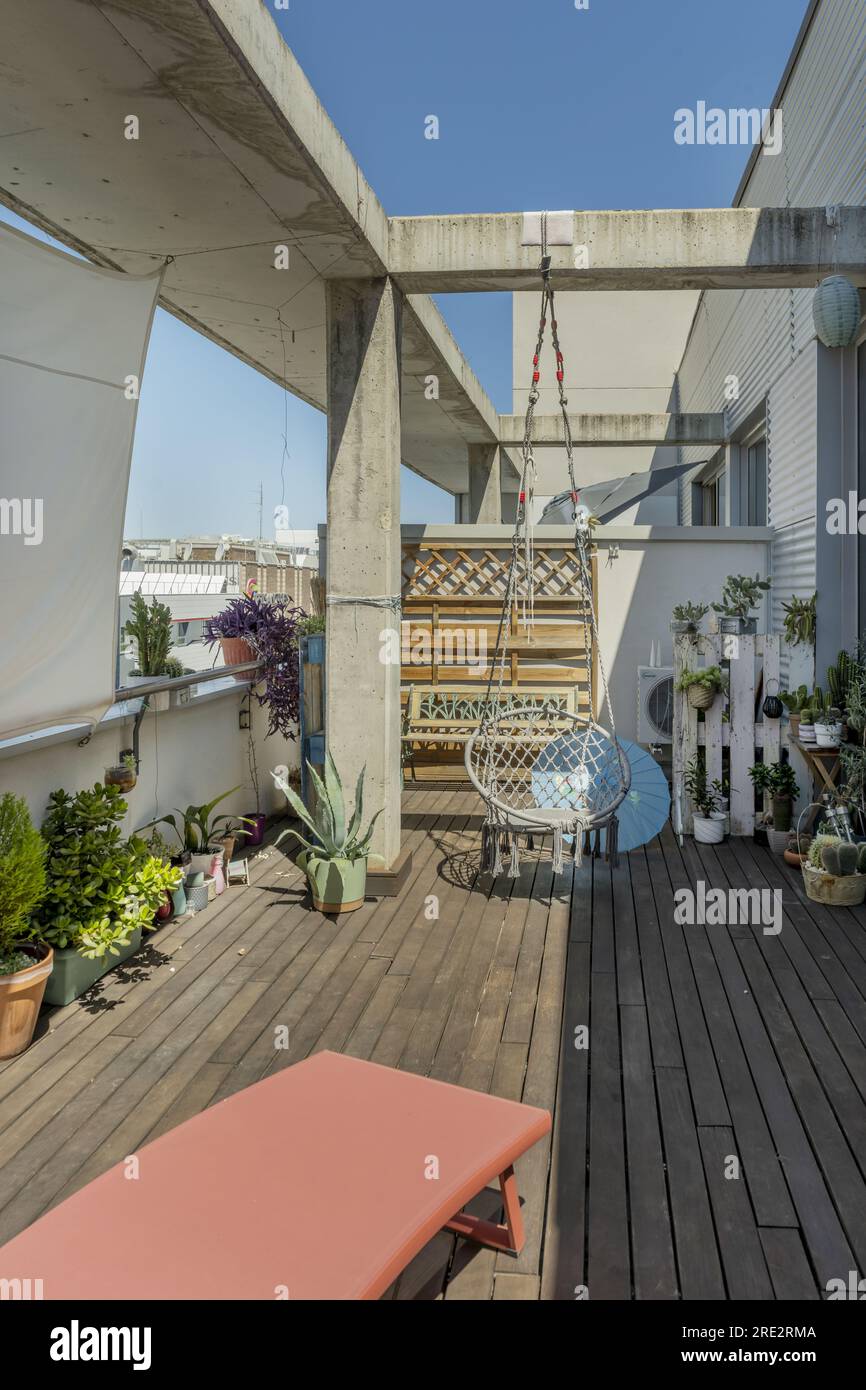 Urbane Penthouse-Terrasse mit Akazienholzboden, dekoriert mit vielen Pflanzen, Betonstruktur, orangefarbener Hängematte, orientalischem Regenschirm mit blauer Tapete Stockfoto