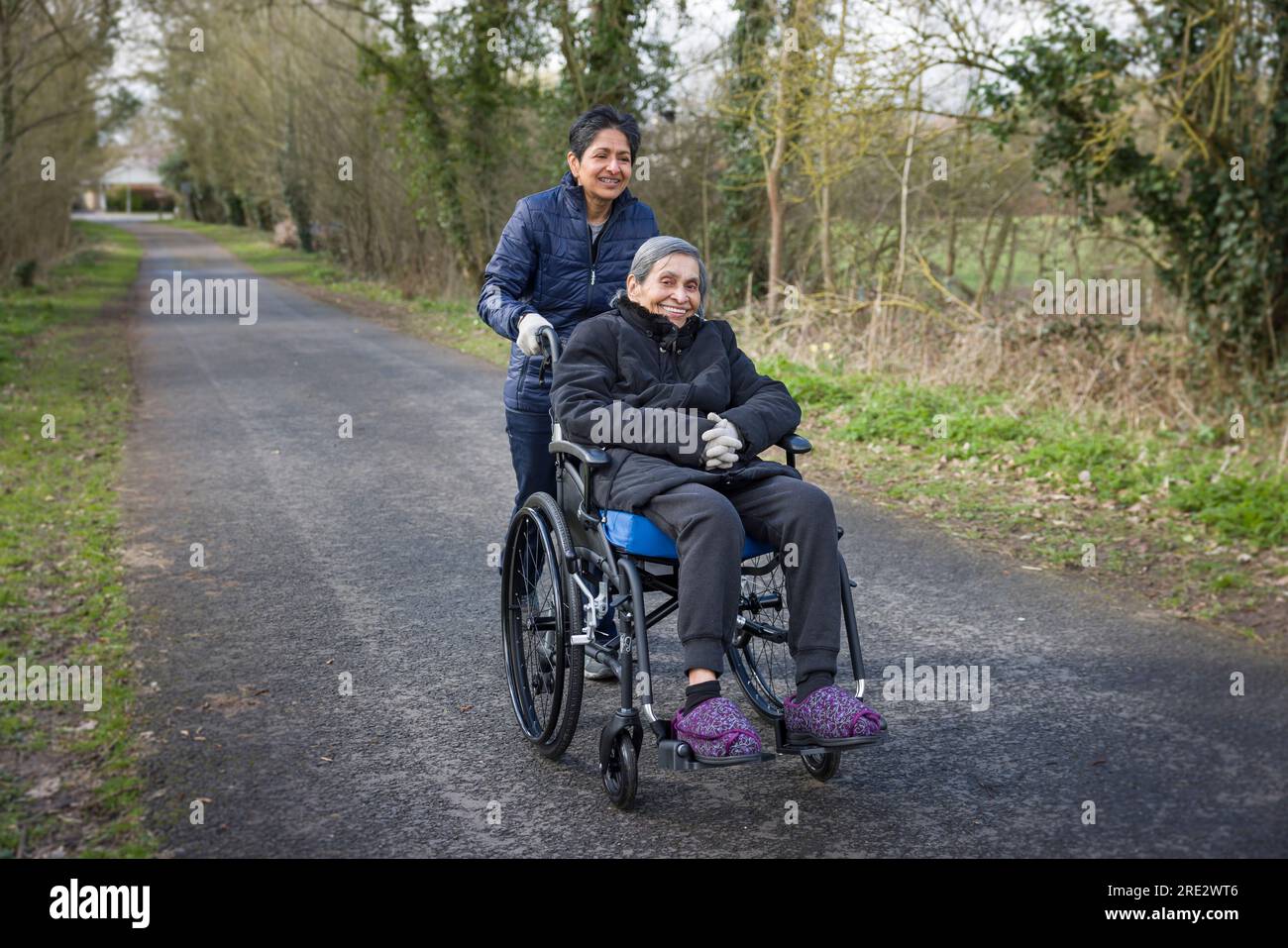 Eine asiatische indische Frau schubst ihre ältere Mutter im Rollstuhl im Winter in Großbritannien nach draußen. Stellen Sie möglicherweise auch einen Betreuer, Fürsorge in der Gemeinde dar Stockfoto