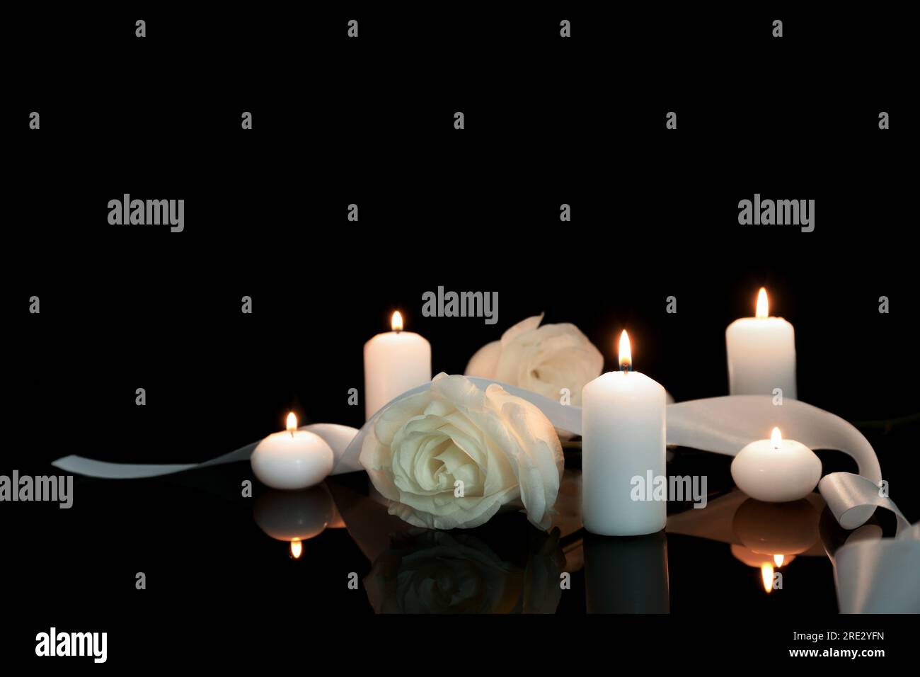 Weiße Rosen und brennende Kerzen auf schwarzer Spiegeloberfläche bei Dunkelheit, Platz für Text. Begräbnissymbole Stockfoto