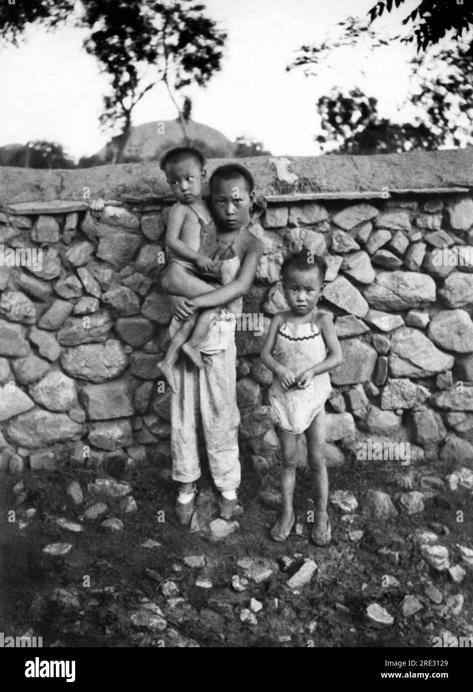 China: ca. 1925 drei Kinder stehen vor einer Steinmauer in einem kleinen Landdorf im ländlichen China. Stockfoto