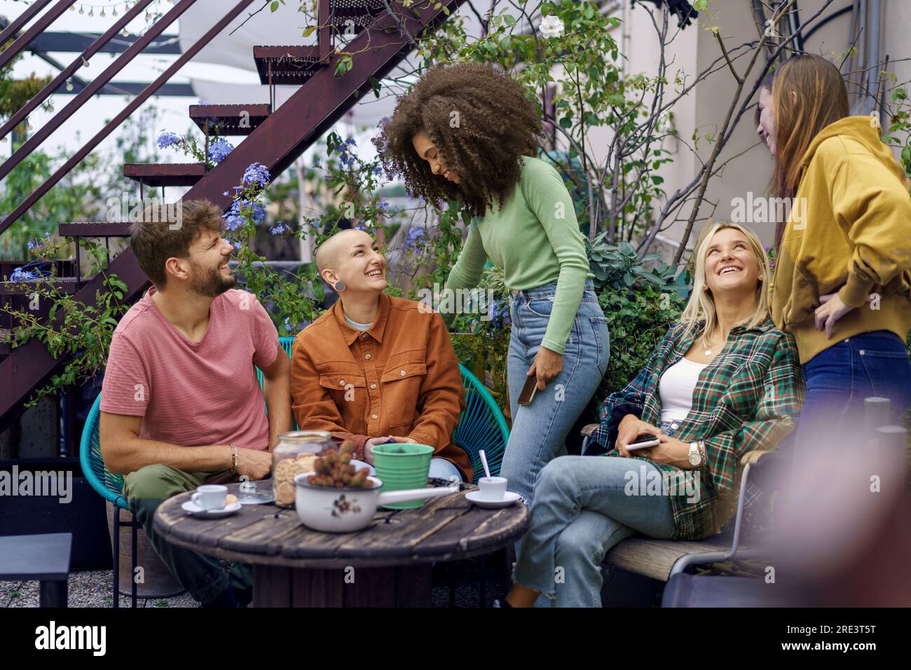 Junge multikulturelle Studenten genießen Espresso und Kekse, interagieren mit einem Lächeln auf einer mit Pflanzen gefüllten Terrasse, mit einigen auf ihren Telefonen. Stockfoto