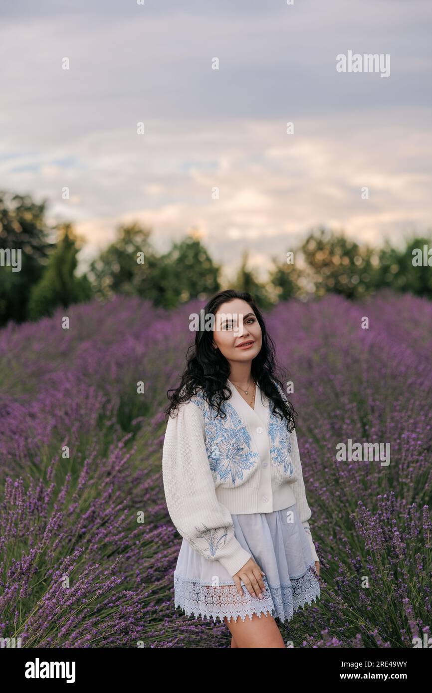 Junge brünette Frau, die spaziert und sich unter blühenden Lavendelreihen vor dem Hintergrund des Sonnenuntergangs amüsiert. Stockfoto