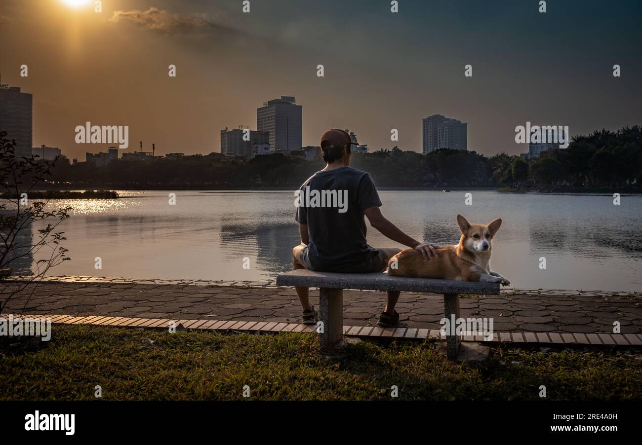 Ein vietnamesischer Mann sitzt mit seinem Corgi-Hund auf einer Bank, während die Sonne über dem See im Reunification Park in Hanoi, Vietnam untergeht. Stockfoto