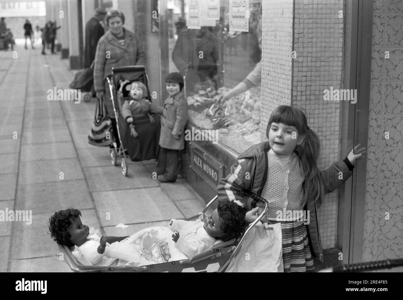 Ein junges weißes Mädchen spielt mit zwei schwarzen Kinderpuppen in einem Kinderwagen Glasgow. Schottland UK. Sie wartet vor einem Laden, während ihre Mutter drinnen einkauft. 1979 HOMER SYKES Stockfoto