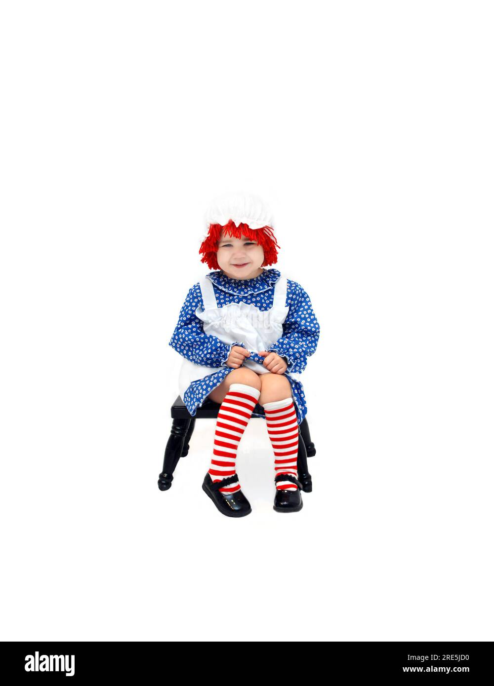 Das süße kleine Mädchen ist als Stoffpuppe verkleidet. Sie grinst schamlos und sitzt auf einem schwarzen Melkhocker. Stockfoto