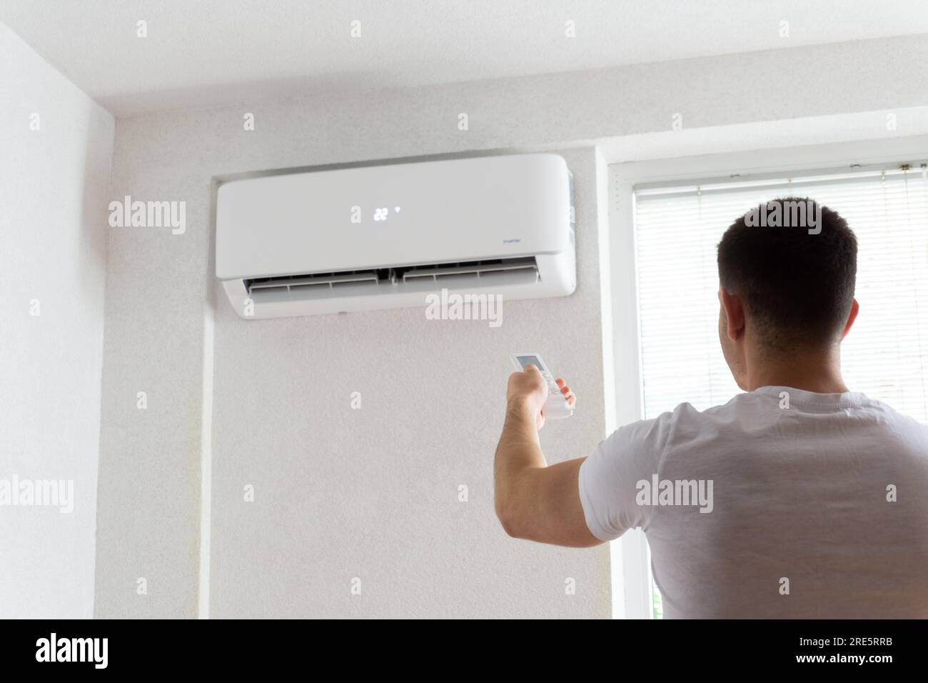 Der junge Mann schaltet die Klimaanlage ein. Hübscher junger Mann, der die Klimaanlage mit Fernbedienung anmacht. Steigende Temperaturen, heißes Wetter. Stockfoto