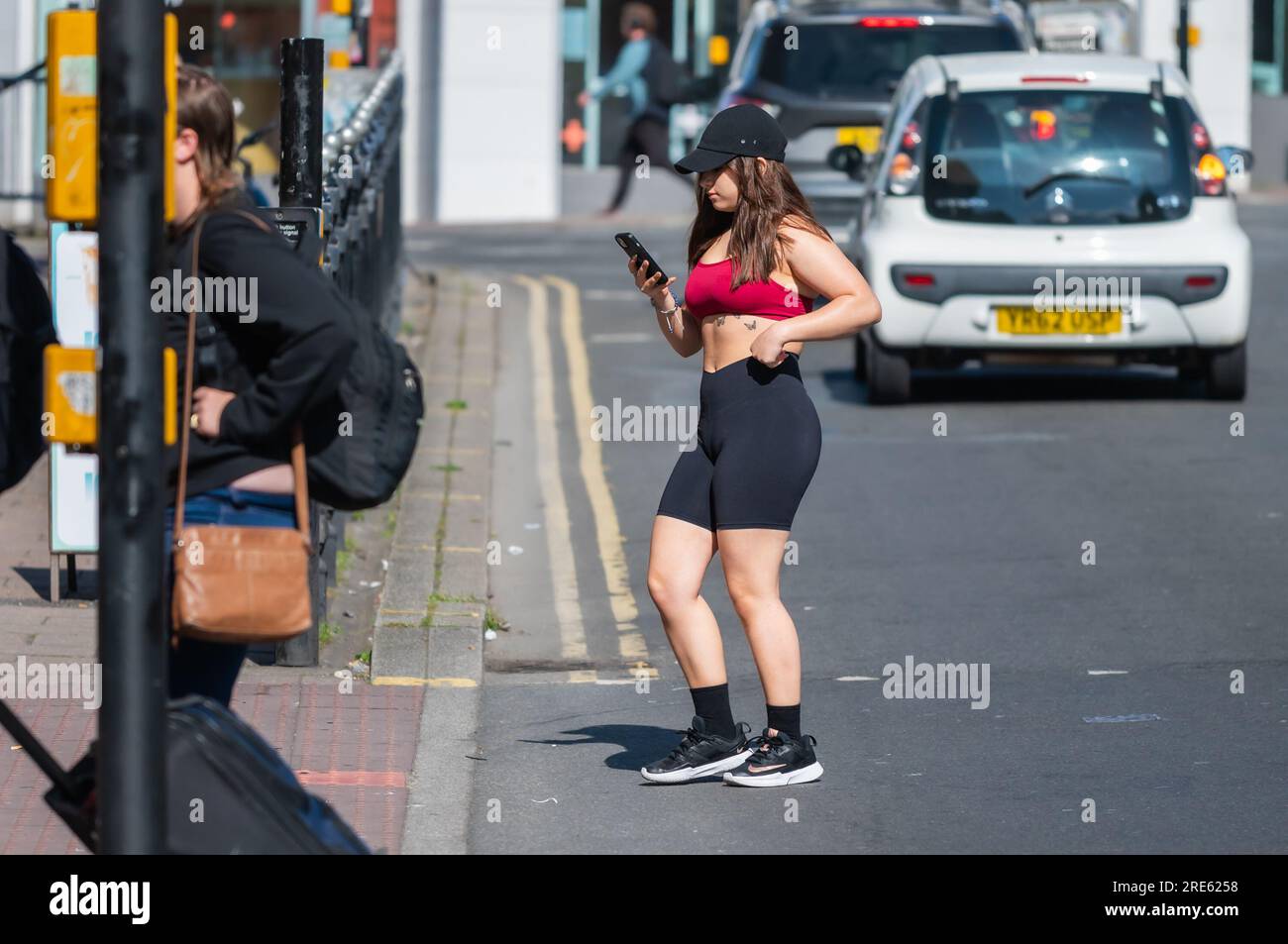 Junge Frau, die für den Sommer gekleidet ist und auf einer Straße steht, mit einem Mobiltelefon, Smartphone oder Mobiltelefon in England, Großbritannien. Stockfoto