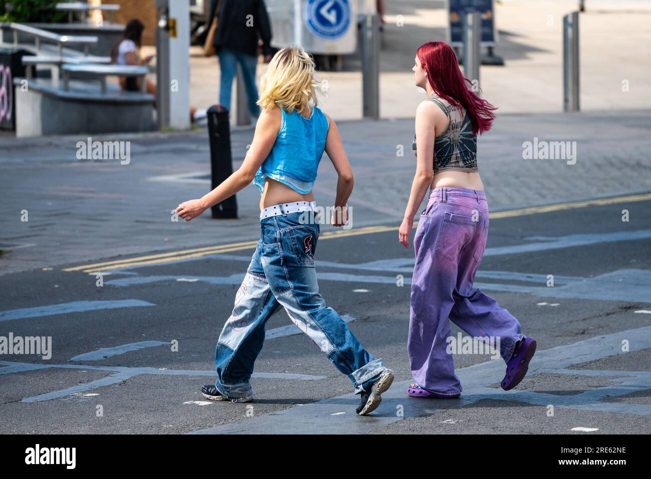 Zwei junge, schlanke Frauen, Mädchen oder Teenager, die in Jeans gekleidet sind, über eine Straße gehen und in Summer, Großbritannien, einkaufen gehen. Freunde in Geschäften. Stockfoto