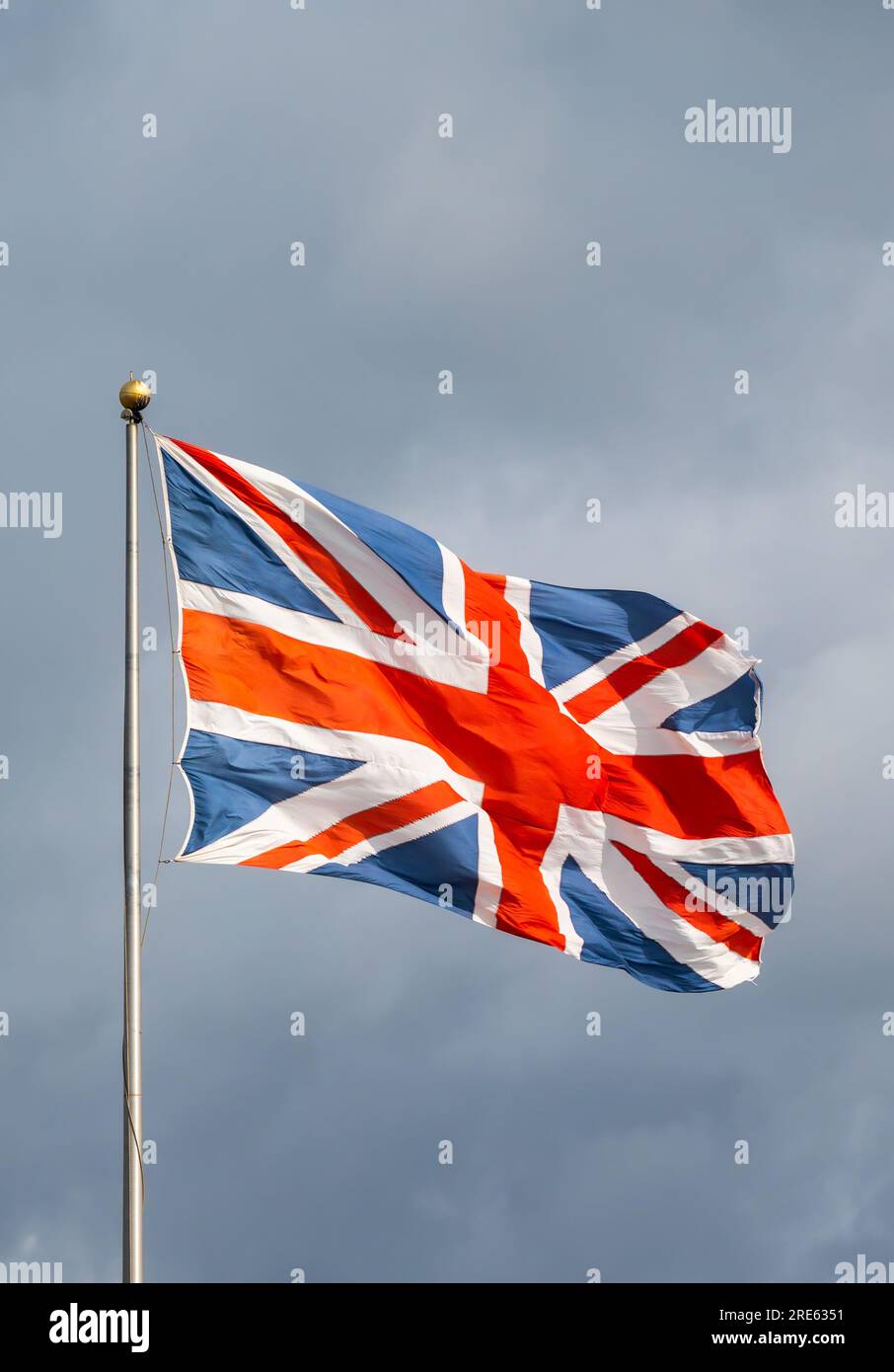 Union Jack, Flagge des Vereinigten Königreichs Großbritannien und Nordirland, fliegt im starken Wind auf einem Stab gegen bewölkten Himmel. Stockfoto