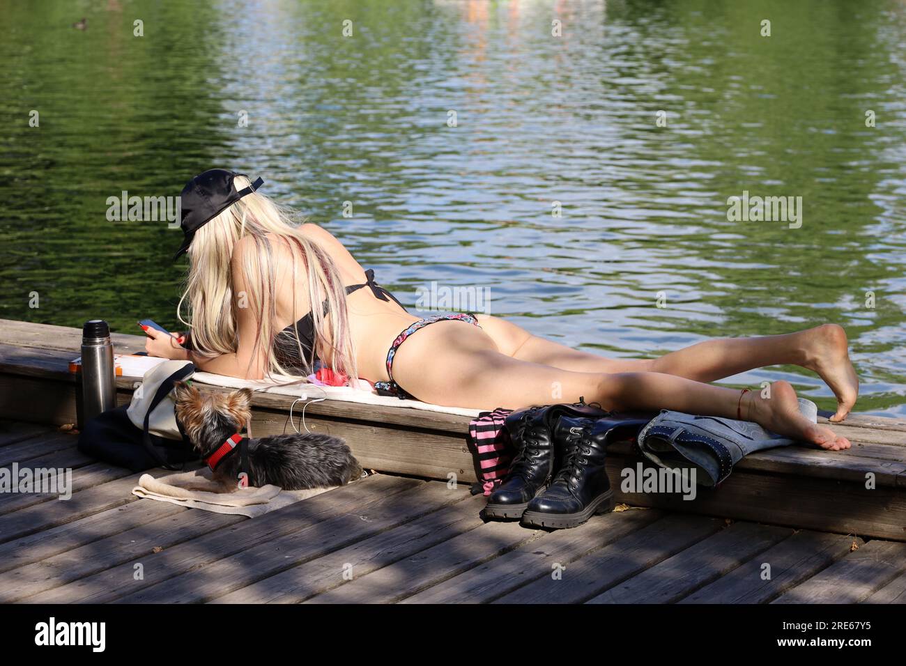 Mädchen im Bikini, Sonnenbaden mit Smartphone, das auf dem Bauch am Ufer des Sees liegt, mit Hund, Sommerurlaub in der Stadt Stockfoto