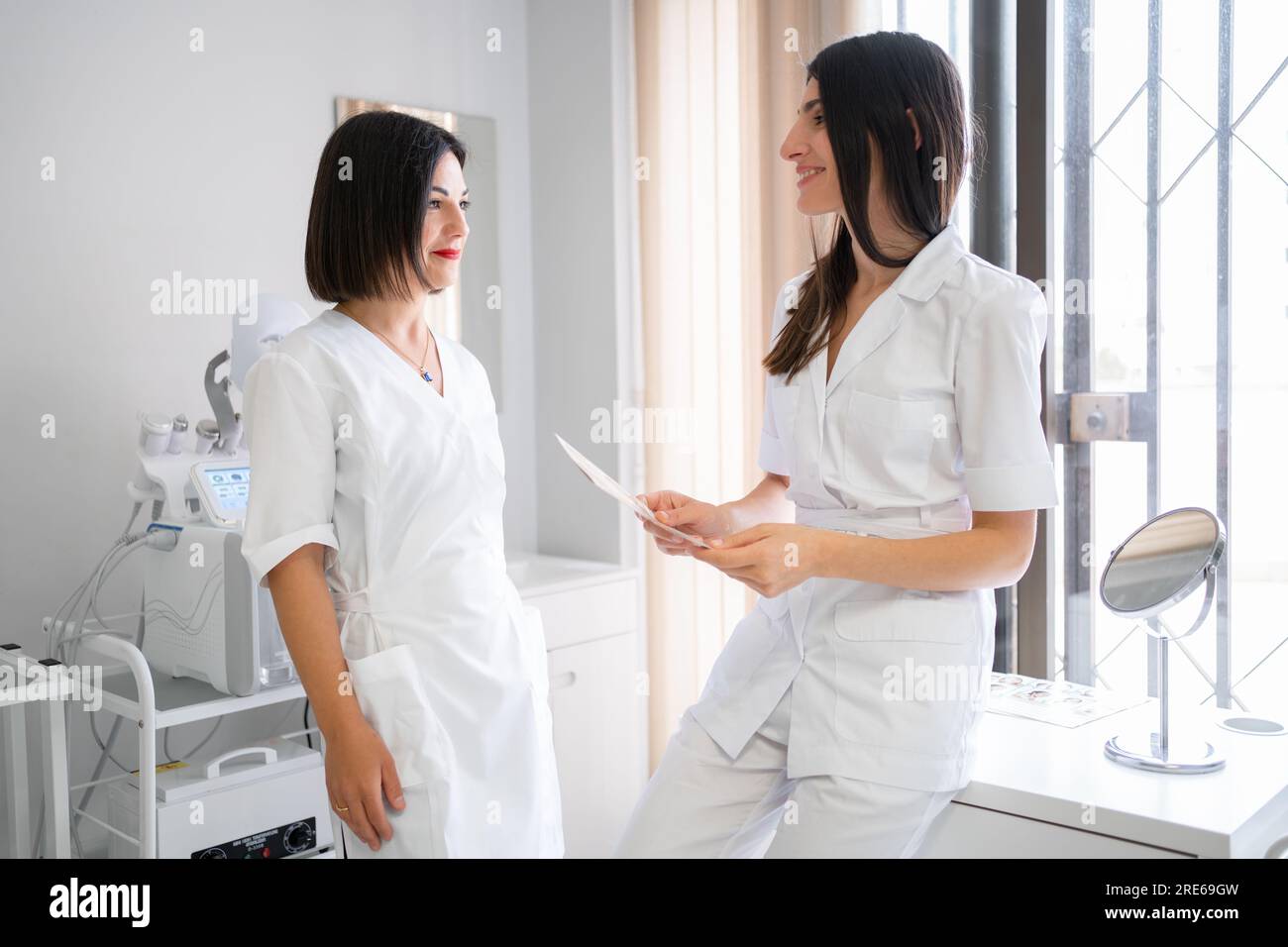 Zwei Ärztinnen stehen in der medizinischen Klinik und diskutieren die Ergebnisse der Patientenuntersuchungen. Sie sehen sich an und lächeln. Spezialisten im Gesundheitswesen führen ein professionelles Gespräch. Stockfoto