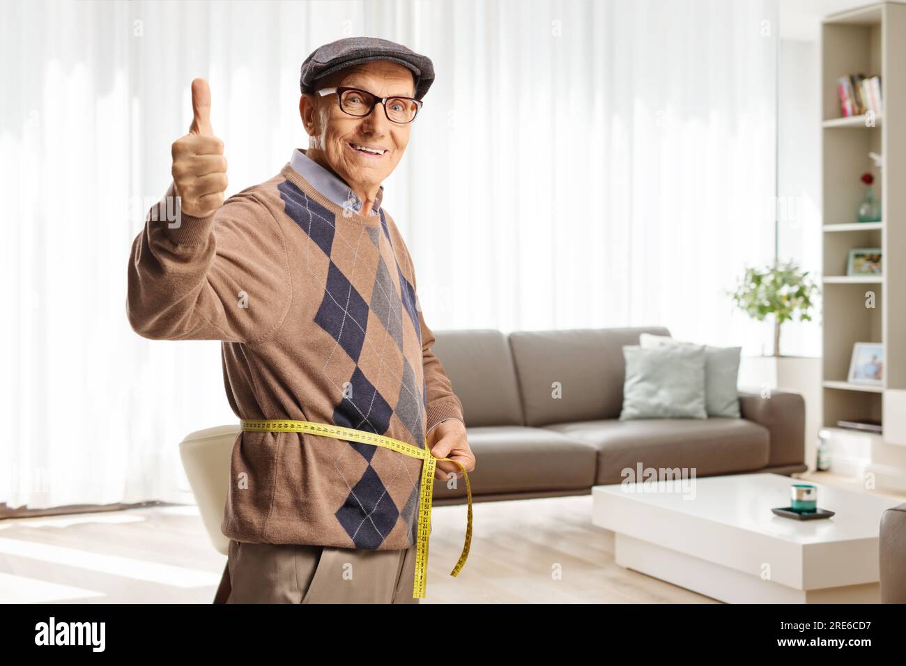 Älterer Mann, der im Wohnzimmer mit einem Klebeband die Taille misst und Daumen nach oben drückt Stockfoto