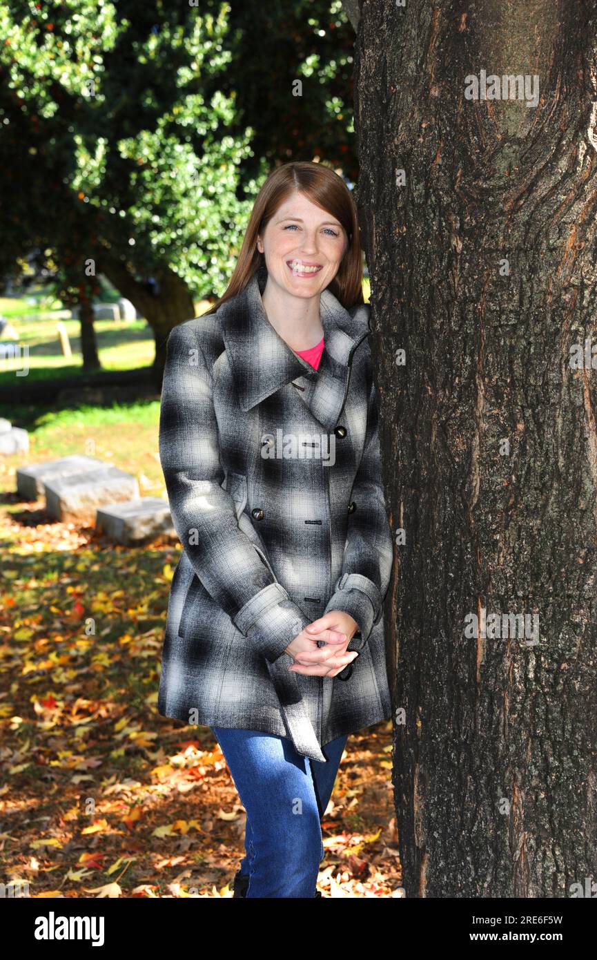 Die junge Frau genießt einen Herbsttag auf dem Elmwood Cemetery in Memphis, Tennessee. Sie lehnt sich an einen Baum und strahlt. Stockfoto