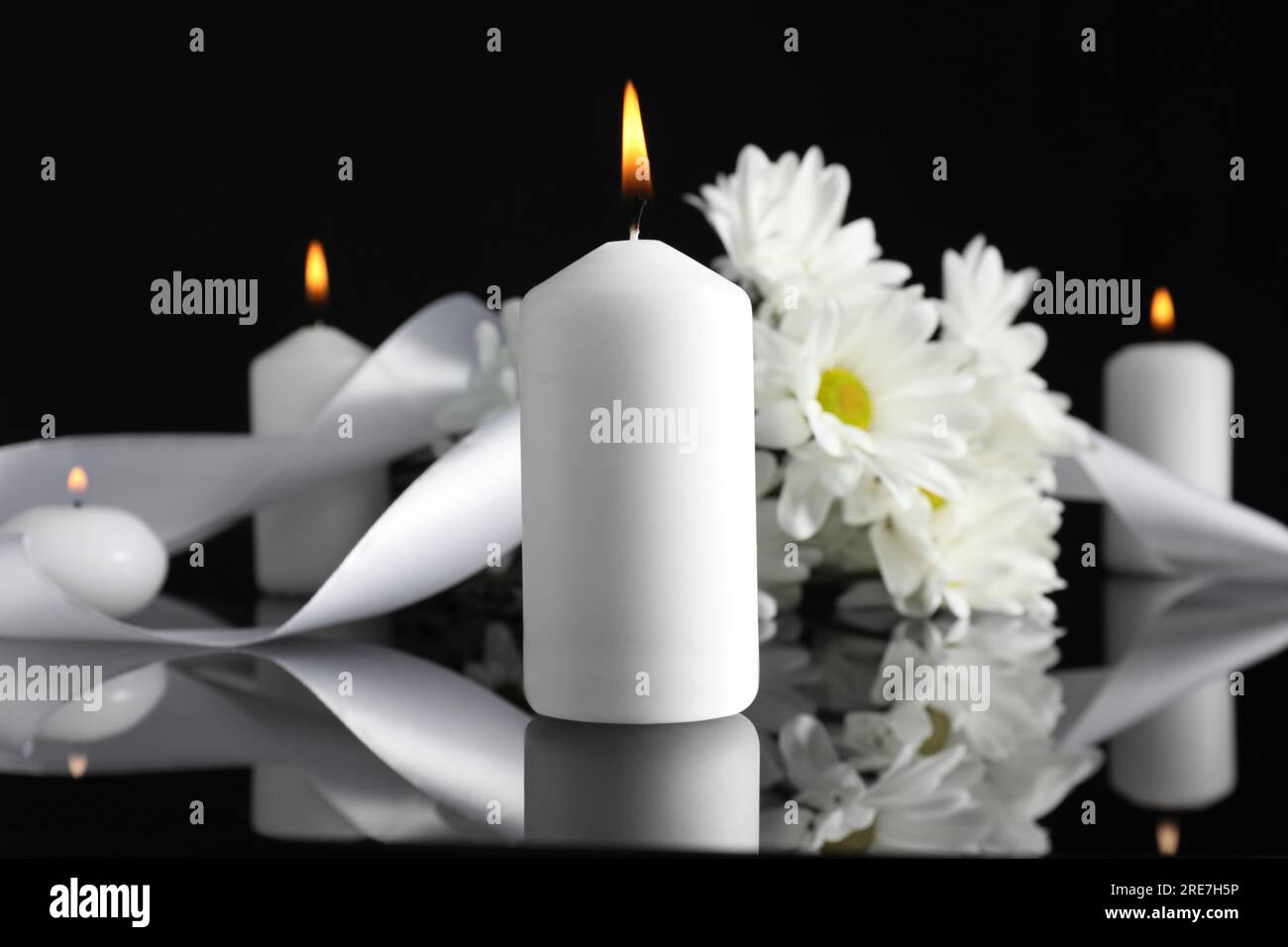 Weiße Chrysanthemen und brennende Kerzen auf schwarzer Spiegeloberfläche in der Dunkelheit, Nahaufnahme. Begräbnissymbole Stockfoto