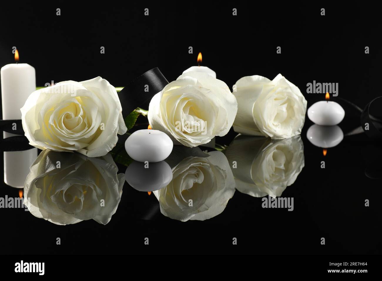 Weiße Rosen und brennende Kerzen auf schwarzer Spiegeloberfläche in der Dunkelheit. Begräbnissymbole Stockfoto