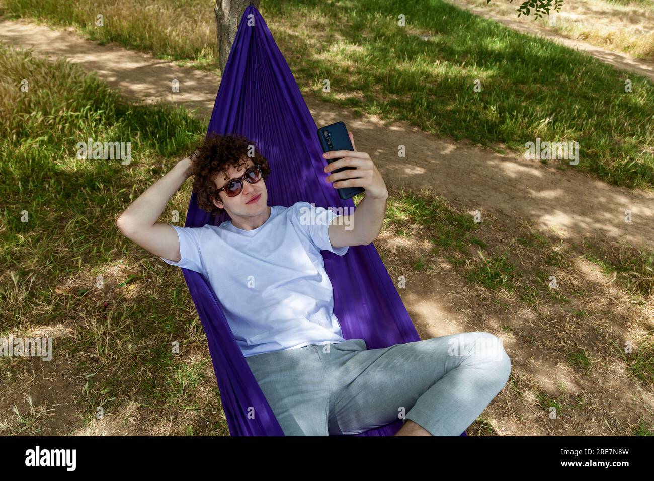 Ein junger Mann macht ein Selfie am Telefon und entspannt sich in einer lila Hängematte in der Natur, genießt die Stille. Stockfoto