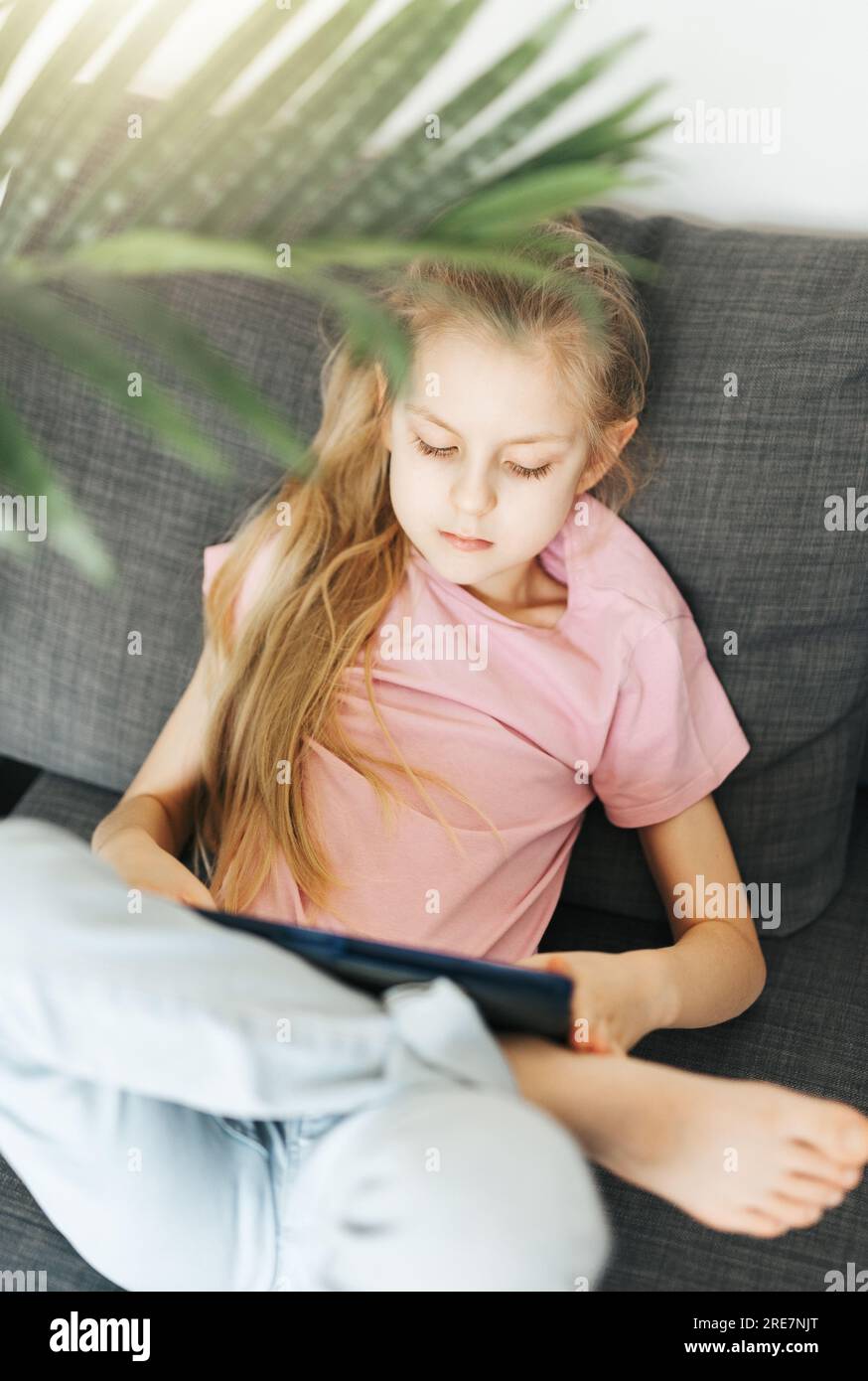 Ein kleines Mädchen, das auf der Couch liegt, mit einem elektronischen Tablet. Einsatz von drahtlosen Geräten für den Bildungsbereich. Stockfoto