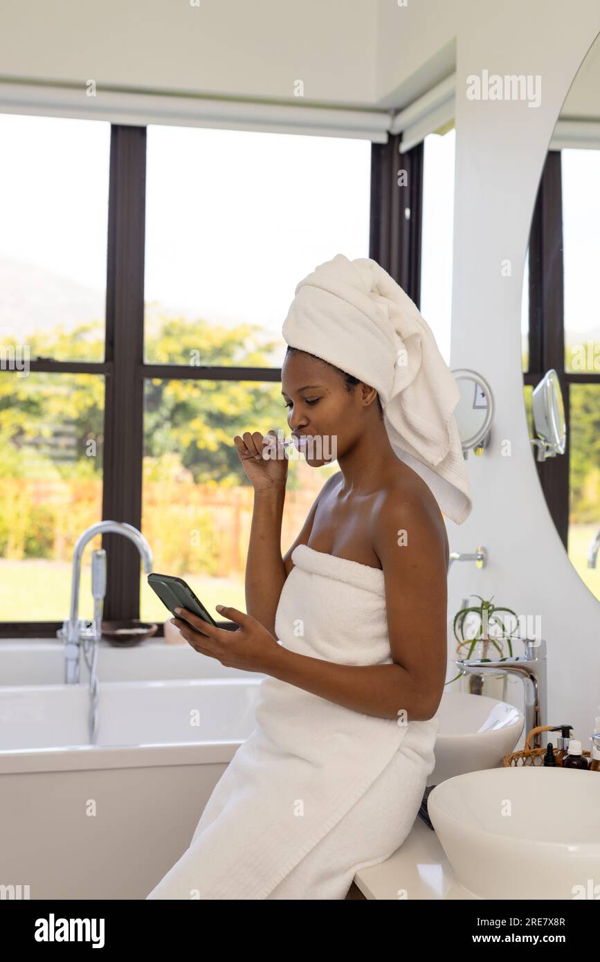 Eine afroamerikanische Frau, die ein Handtuch am Kopf trägt, Zähne putzt und im Badezimmer ein Smartphone benutzt Stockfoto