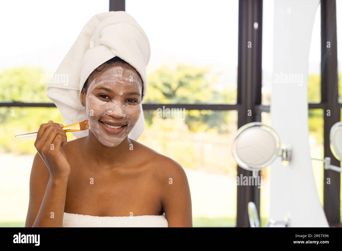 Portrait einer glücklichen afroamerikanischen Frau, die ein Handtuch auf dem Kopf trägt und eine Gesichtsmaske im Badezimmer aufbringt Stockfoto