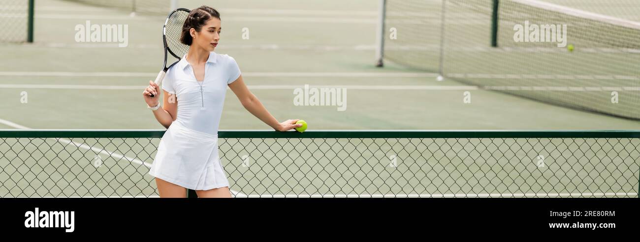 Sportlerin in weißer Sportbekleidung hält Ball und Schläger, während sie neben Tennisnetz und Banner steht Stockfoto