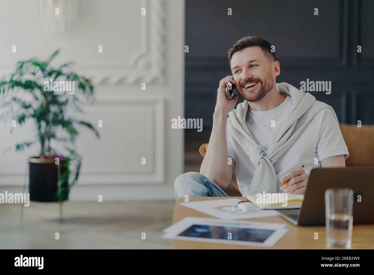 Gutaussehender Geschäftsmann/Freiberufler, ungezwungene Kleidung, Telefonieren am Heimbüro mit Laptop, weißes T-Shirt und Pullover über den Schultern, klein Stockfoto