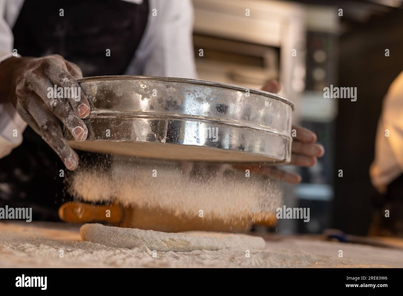 afroamerikanischer Bäcker trägt Schürze in der Bäckerei und gießt Mehl auf Teig Stockfoto