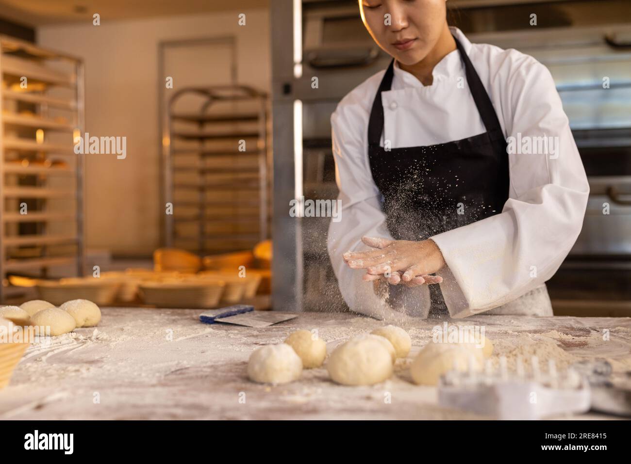 Fokussierte asiatische Bäckerin in einer Bäckerei, die eine Schürze trägt und Teig für Brötchen bildet Stockfoto