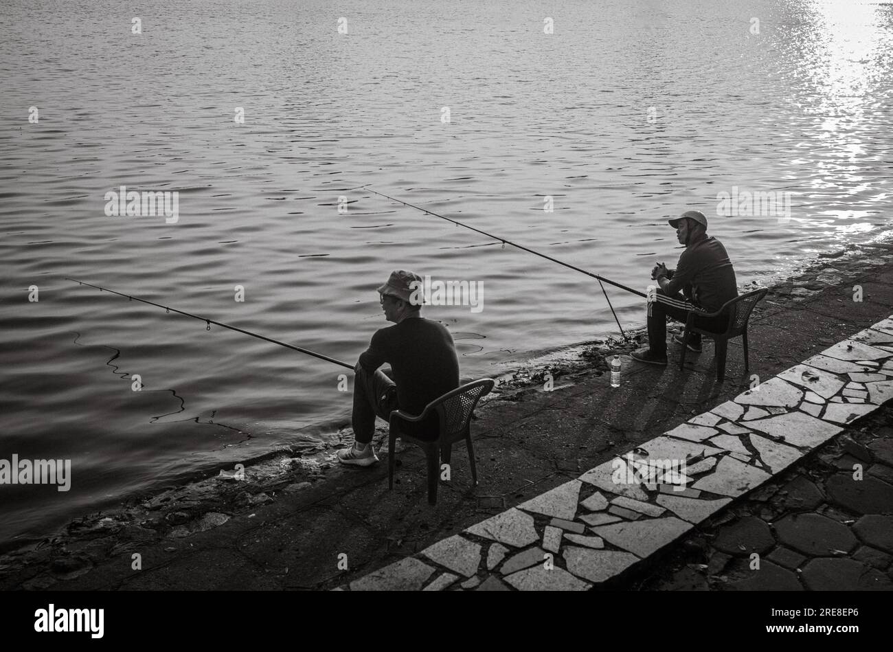 Zwei vietnamesische Männer sitzen im Reunification Park im Zentrum von Hanoi, Vietnam, und fischen bei Sonnenuntergang im See. Stockfoto