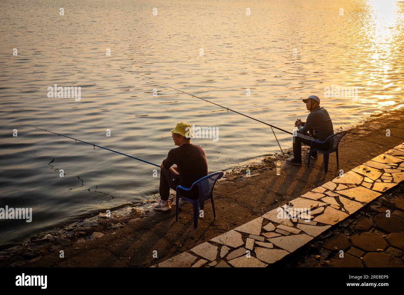 Zwei vietnamesische Männer sitzen im Reunification Park im Zentrum von Hanoi, Vietnam, und fischen bei Sonnenuntergang im See. Stockfoto