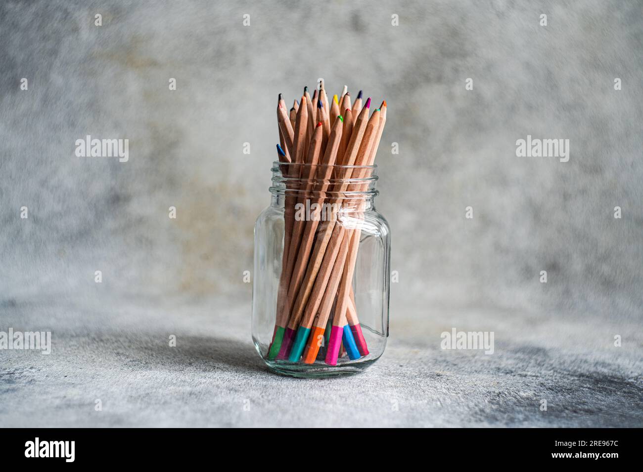 Sammlung von bunten Bleistiften, die in einem Glasbecher gestapelt und vor grauem Hintergrund auf dem Tisch platziert sind Stockfoto